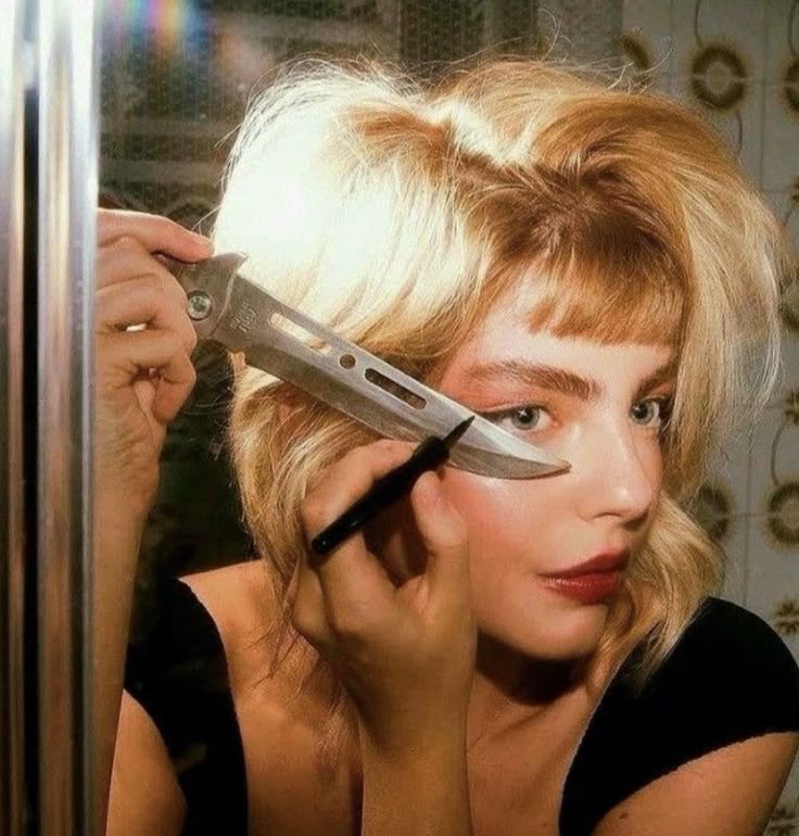 This may contain: a woman holding a knife in front of her face and looking at herself through the mirror